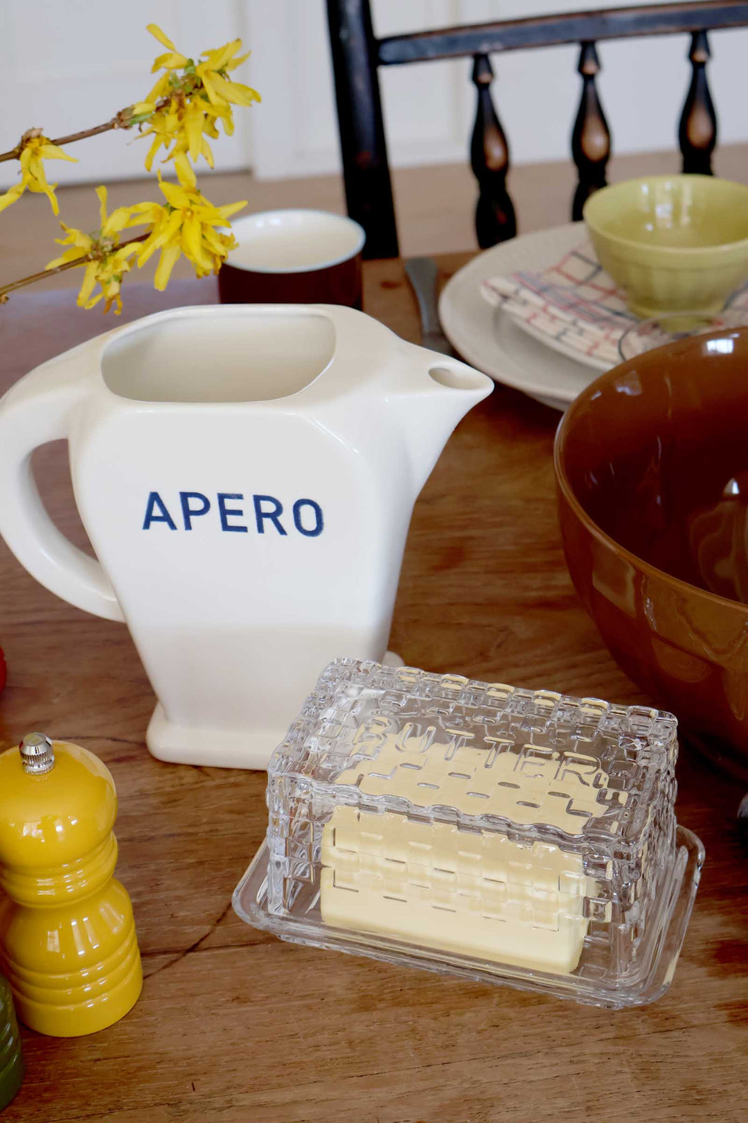 White pitcher with 'APERO' text on a wooden table with yellow flowers and other items.