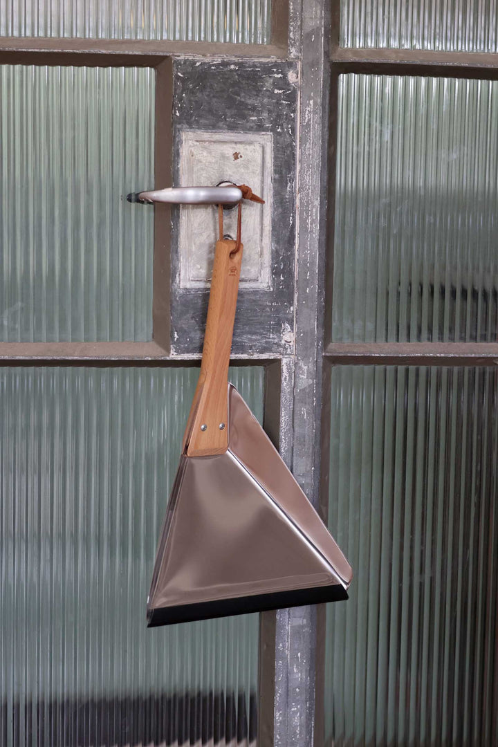 A triangular metal dustpan with a light brown wooden handle hangs from a metal door hook in front of ribbed glass panels.