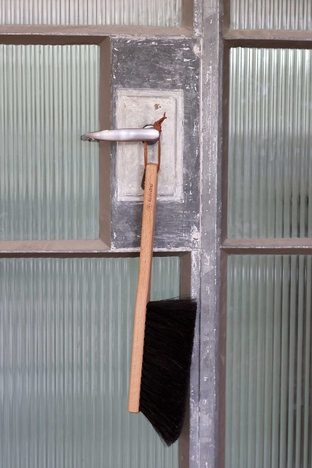 A long-handled black brush with a light wood handle hangs from a weathered metal door handle in front of ribbed frosted glass panels.