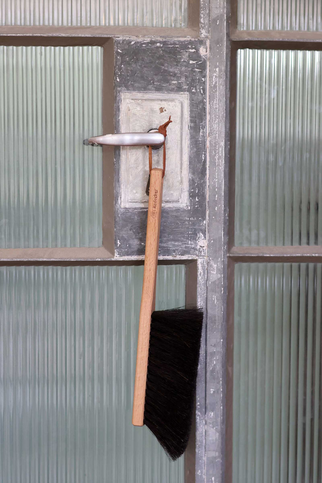 A long-handled black brush with a light wood handle hangs from a weathered metal door handle in front of ribbed frosted glass panels.