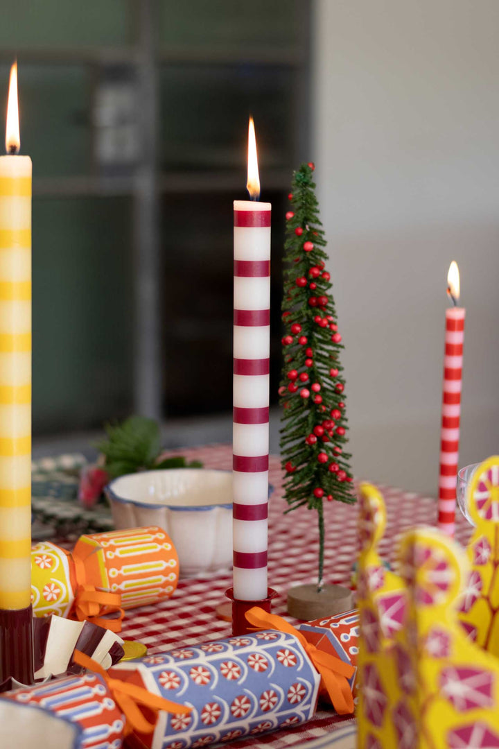 A festive tabletop arrangement with three lit taper candles, including one tall white candle with red stripes, a yellow-and-white striped candle, and smaller patterned crackers, alongside a small decorated Christmas tree in a real indoor setting.