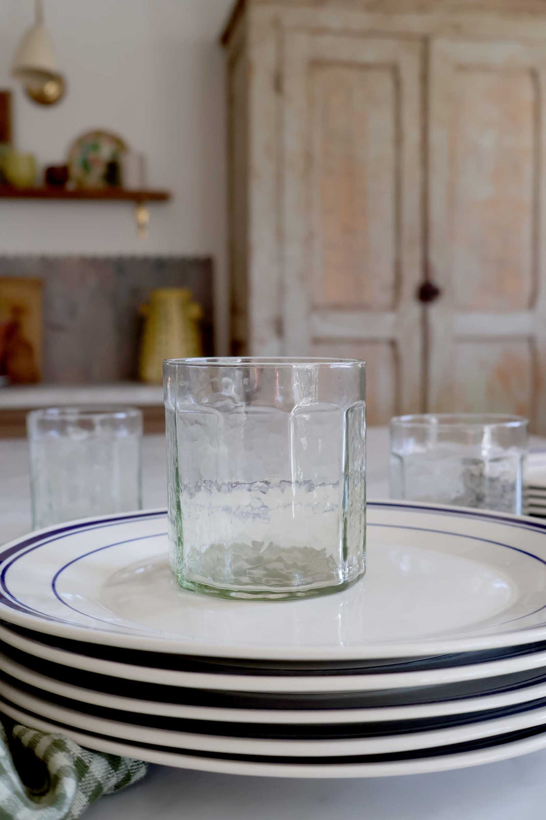 Stack of black-rimmed white plates with a clear glass on a table in a kitchen setting.