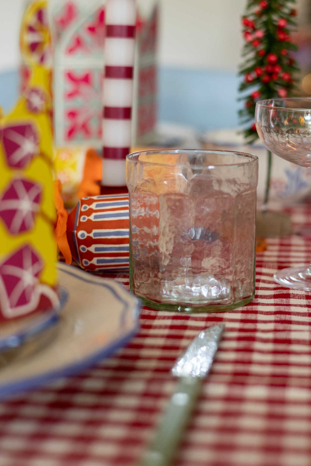 A clear textured glass tumbler on a red and white checkered tablecloth, surrounded by festive holiday decorations including a striped ornament, a small Christmas tree, and patterned plates.