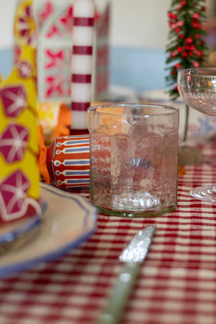 A clear textured glass tumbler on a red and white checkered tablecloth, surrounded by festive holiday decorations including a striped ornament, a small Christmas tree, and patterned plates.