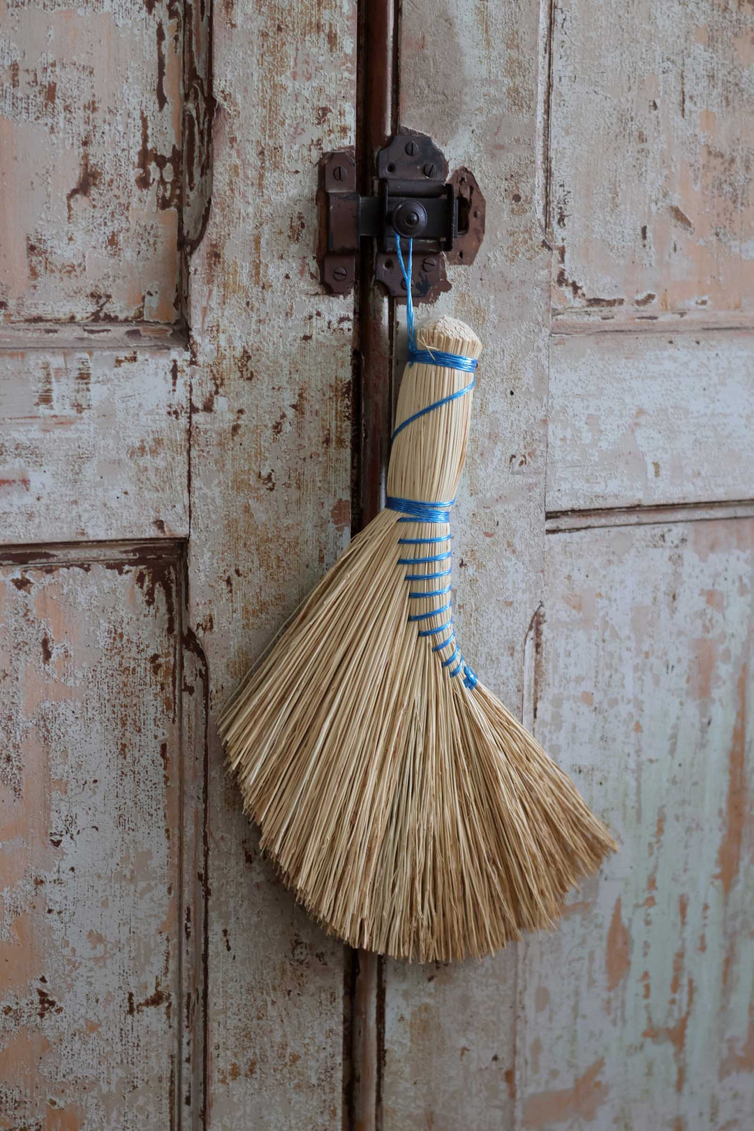 A small traditional straw broom with a blue cord wrap hanging from a rustic metal latch on a weathered, peeling-paint wooden door.