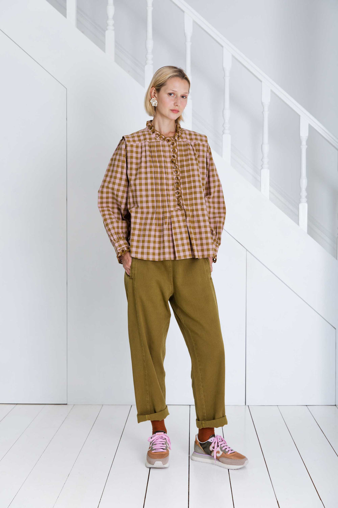 Person wearing a checkered shirt and green pants standing in a minimalistic room with white walls and floor.
