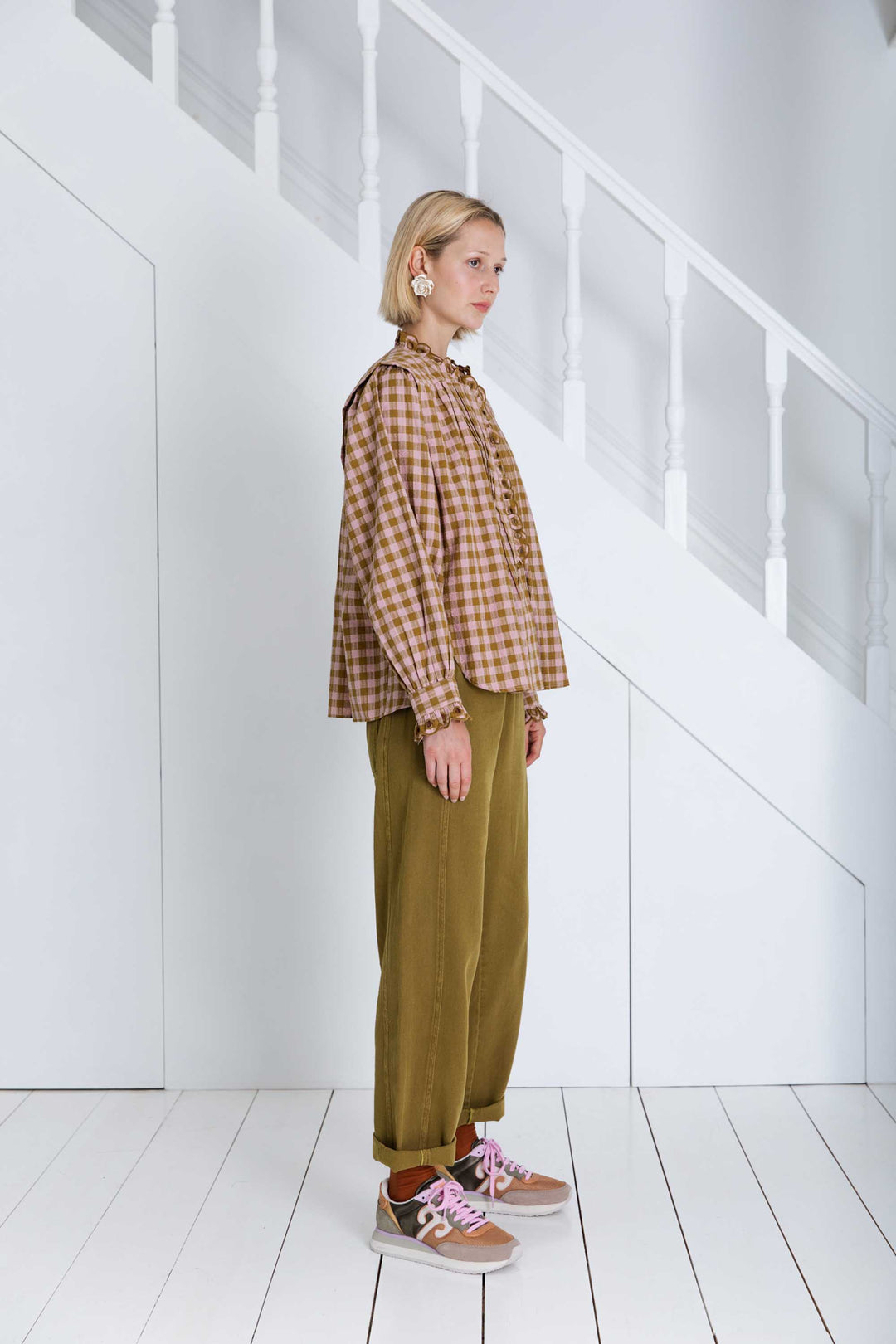 Person wearing a plaid shirt and brown pants standing in a minimalistic room with white walls and floor.