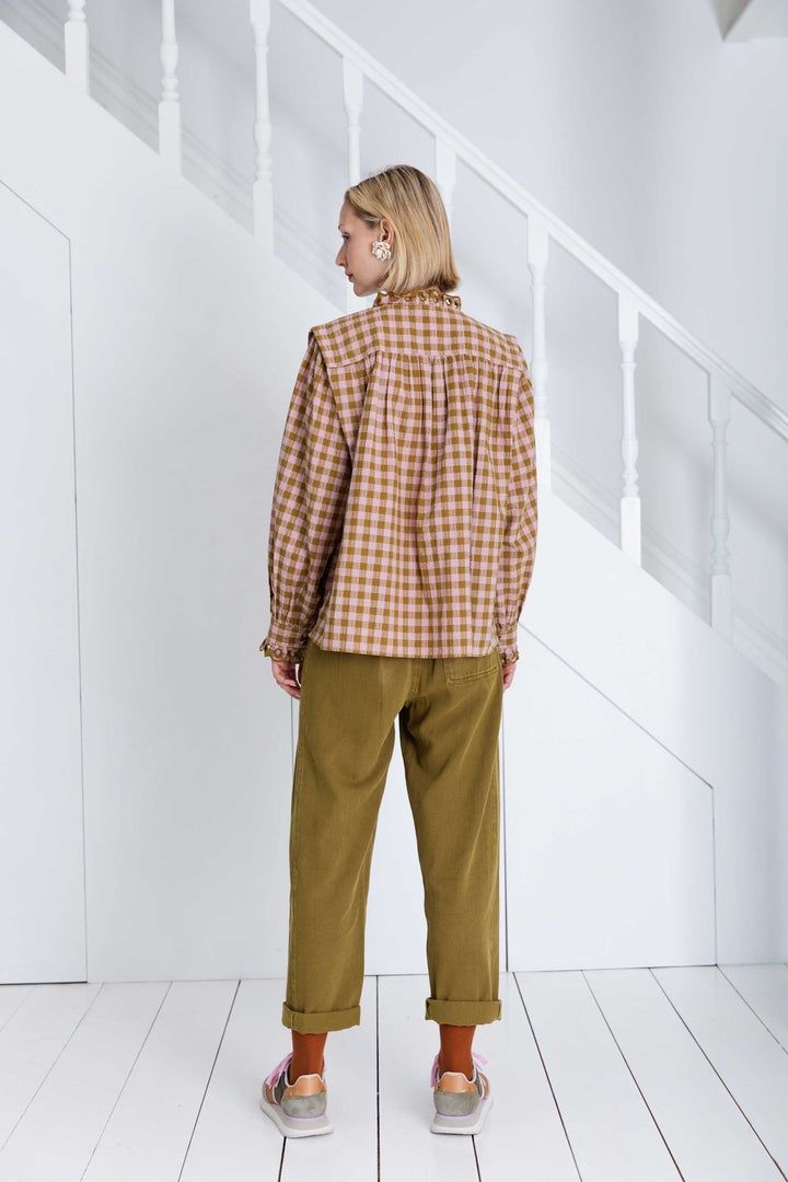 Person wearing a plaid shirt and green pants standing in a minimal white room.