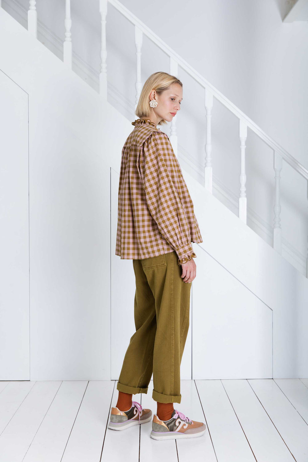Person wearing a plaid shirt and green pants standing in a minimal white room.