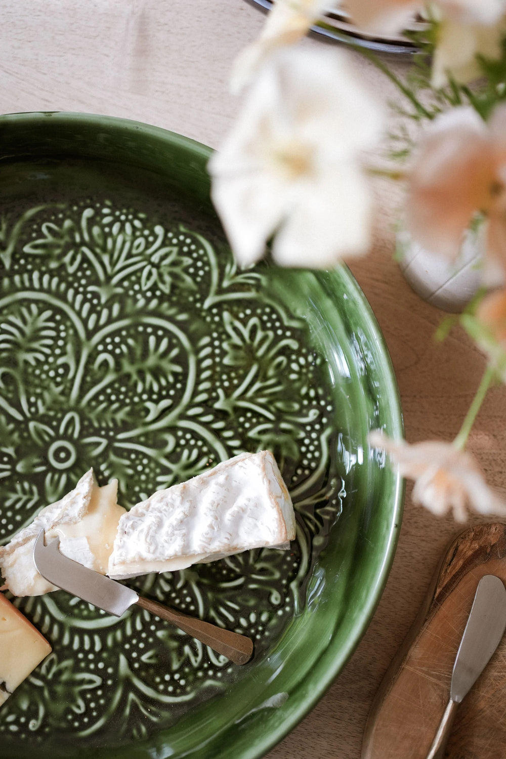Green ceramic plate with cheese and bread on a wooden table with flowers.