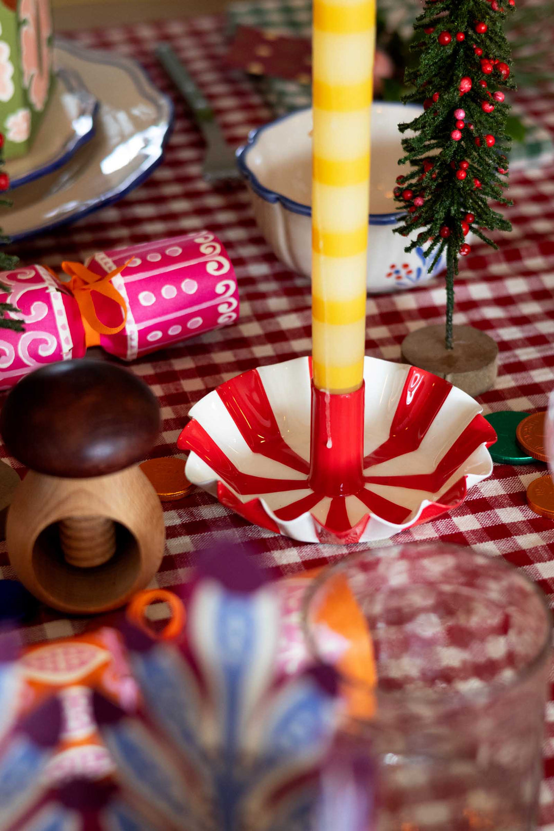 A festive tabletop scene with a red and white striped paper cup holder holding a yellow-and-white striped candle. The setting includes a red-and-white checkered tablecloth, a small Christmas tree, a pink gift-wrapped cracker, and other holiday decorations.