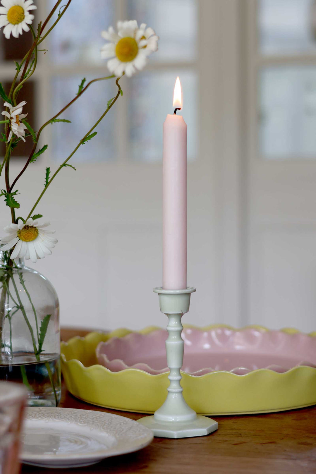 Pink candle in a white holder on a table with flowers and a yellow dish in the background.