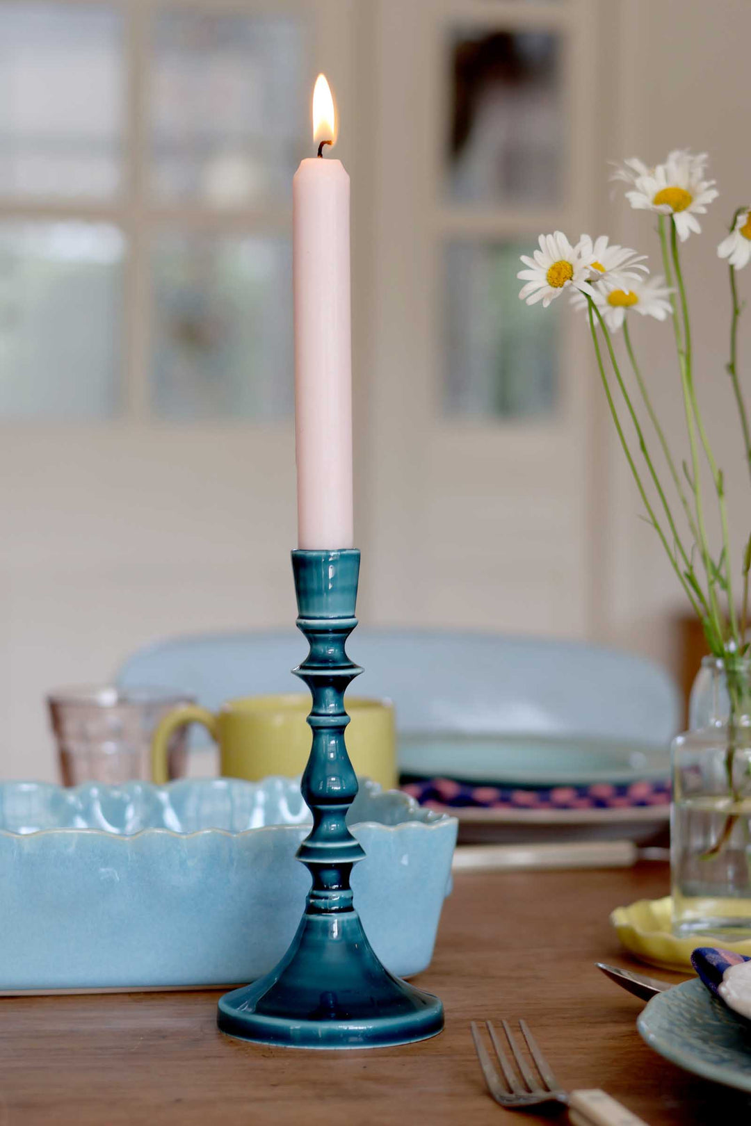 Blue candlestick with a lit pink candle on a dining table with dishes and flowers.