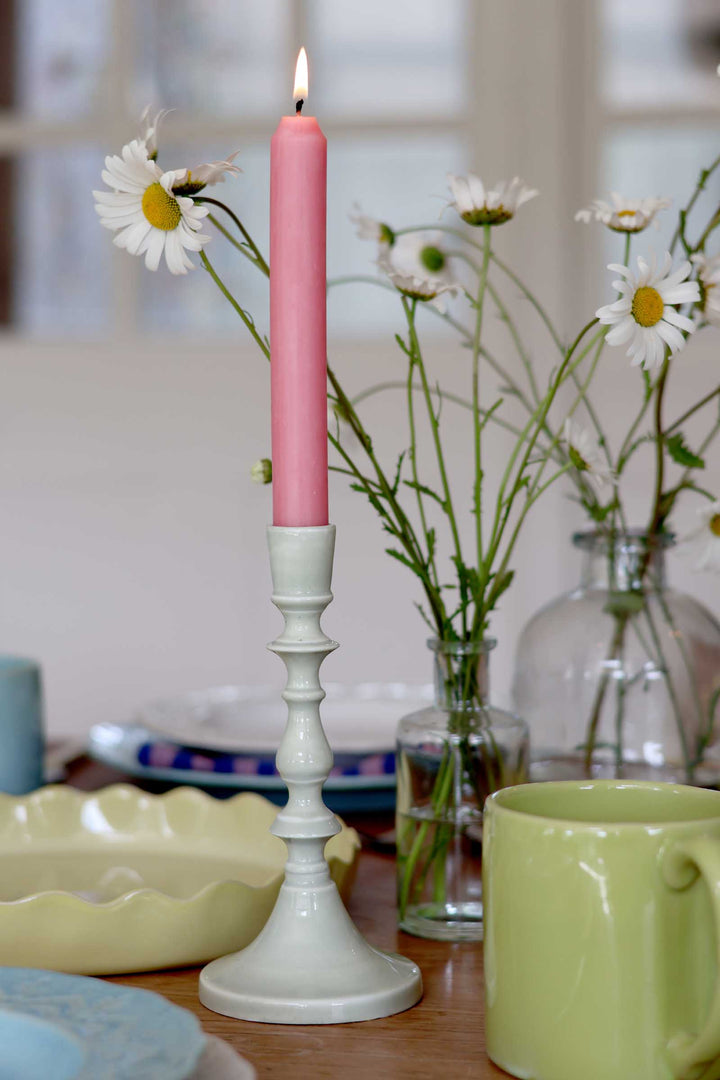 Pink candle in a white holder with flowers and a green mug on a table.