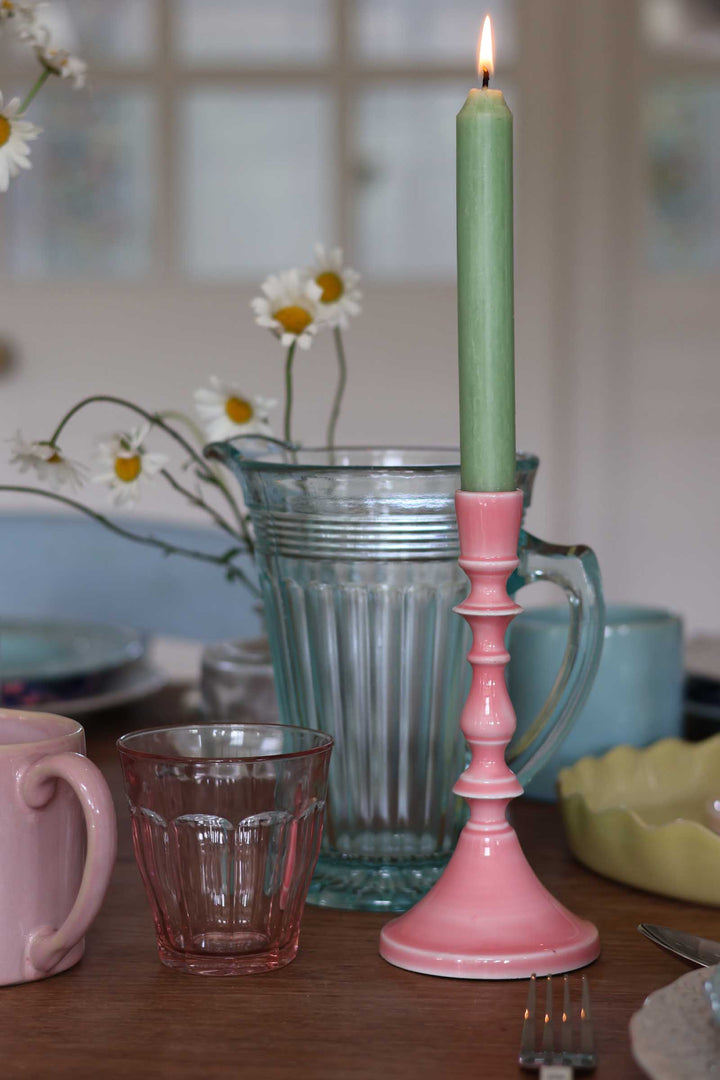 Pink candlestick with a green candle on a table with a vase of flowers and mugs.
