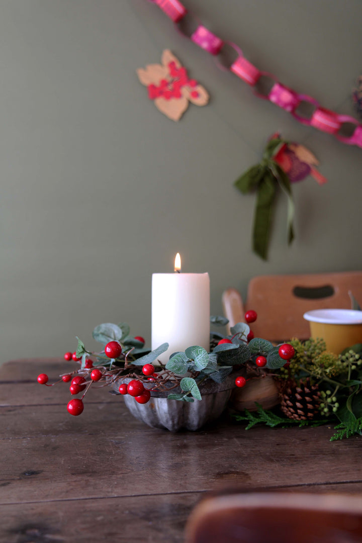 Decorative candle with berries on a wooden table against a neutral wall with festive decorations.