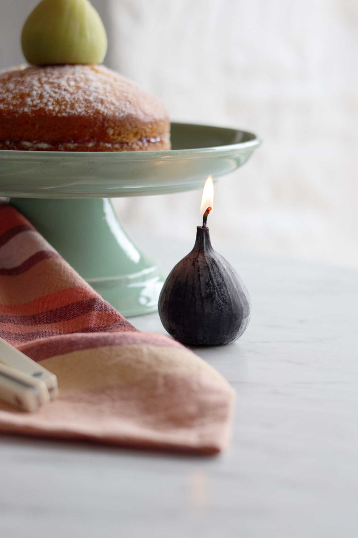 A dark fig-shaped candle with a small flame burning from the top, styled on a light gray tabletop next to a striped napkin and a green cake stand holding a pastry.