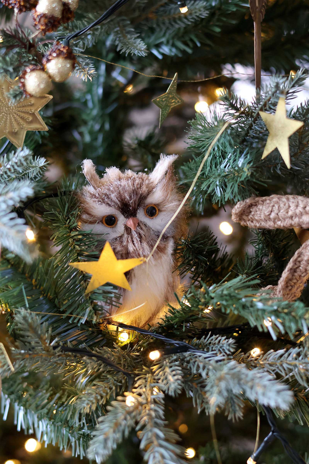 A plush owl Christmas ornament nestled among green tree branches, warm white fairy lights, and gold star decorations on a Christmas tree.