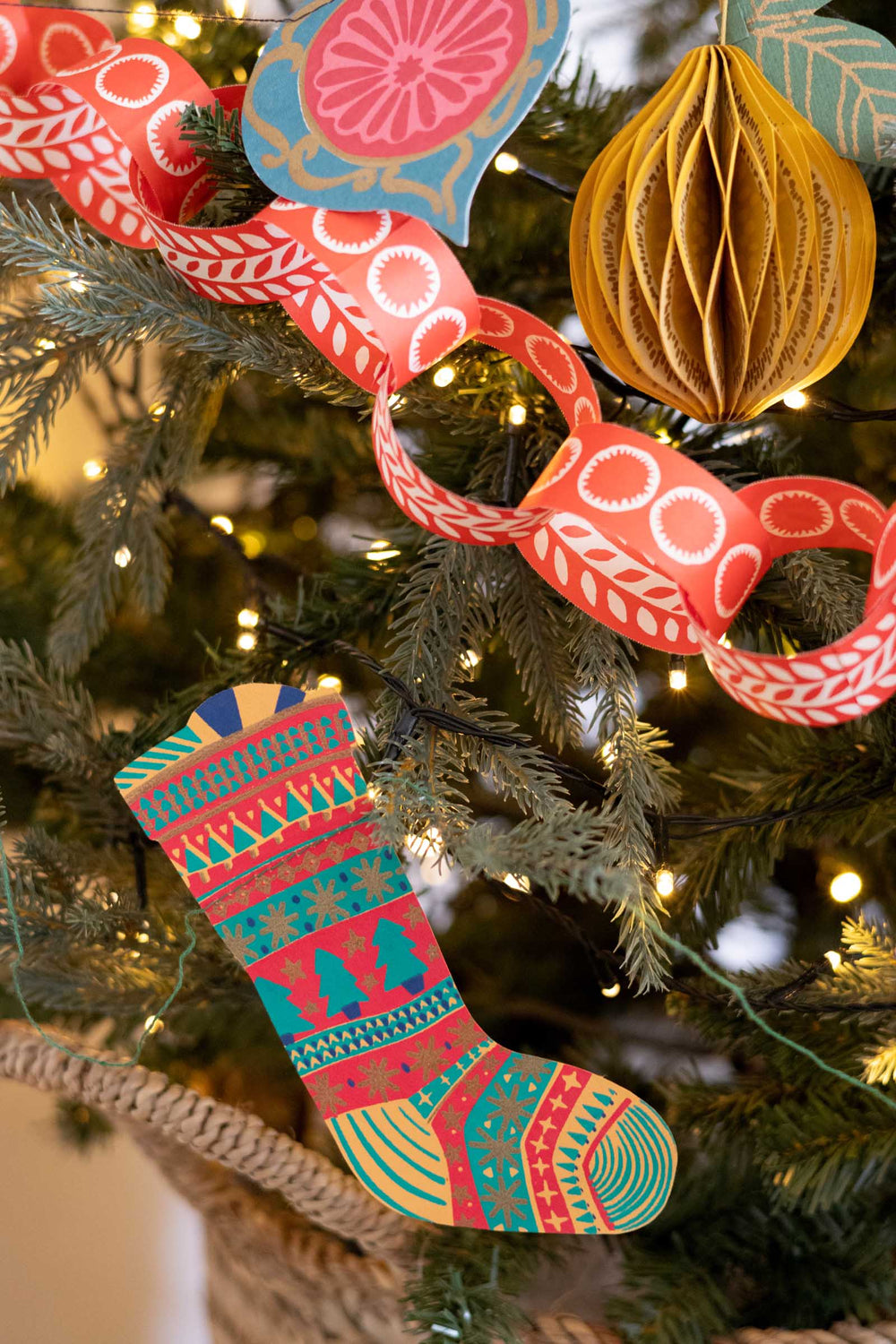 A close-up of a decorated Christmas tree with colorful ornaments, including a patterned Christmas stocking, a red paper chain, a gold paper ball, and a blue citrus slice ornament.