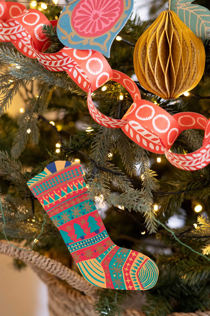 A close-up of a decorated Christmas tree with colorful ornaments, including a patterned Christmas stocking, a red paper chain, a gold paper ball, and a blue citrus slice ornament.