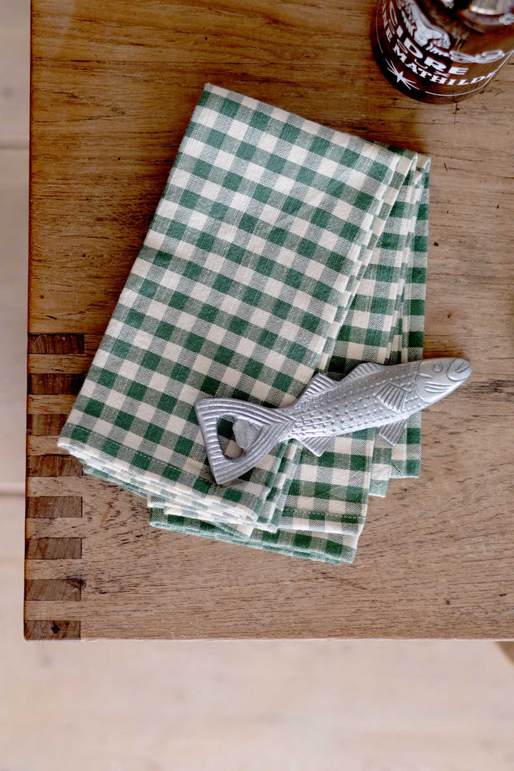 A green and white checkered cloth napkin folded on a wooden table, with a silver fish-shaped bottle opener resting on top.