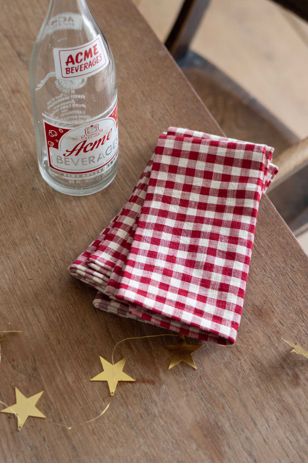 A folded red and white gingham checkered cloth napkin sits on a wooden table next to a vintage clear glass bottle with an Acme Beverage label.