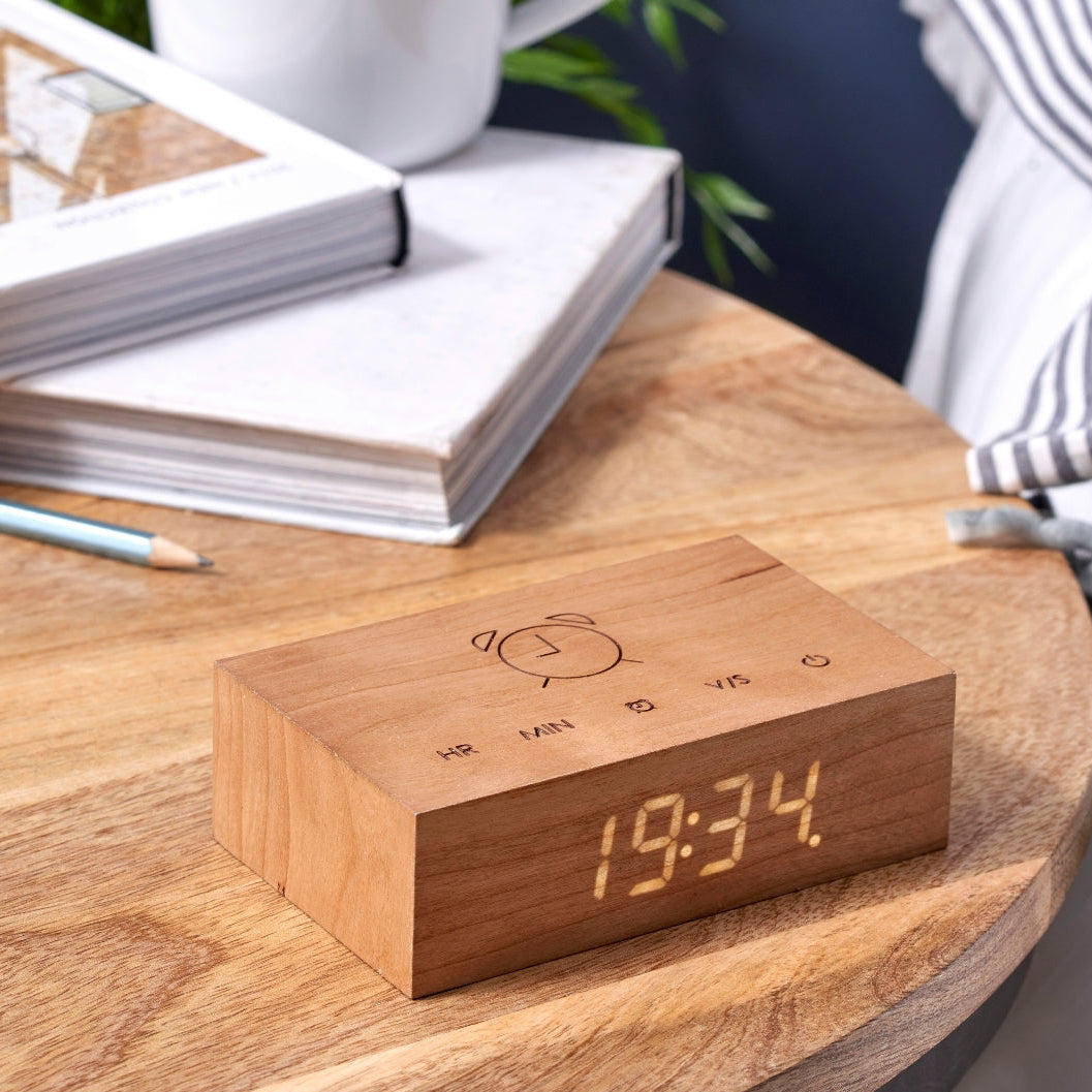 Wooden digital clock on a wooden surface with books and a plant in the background