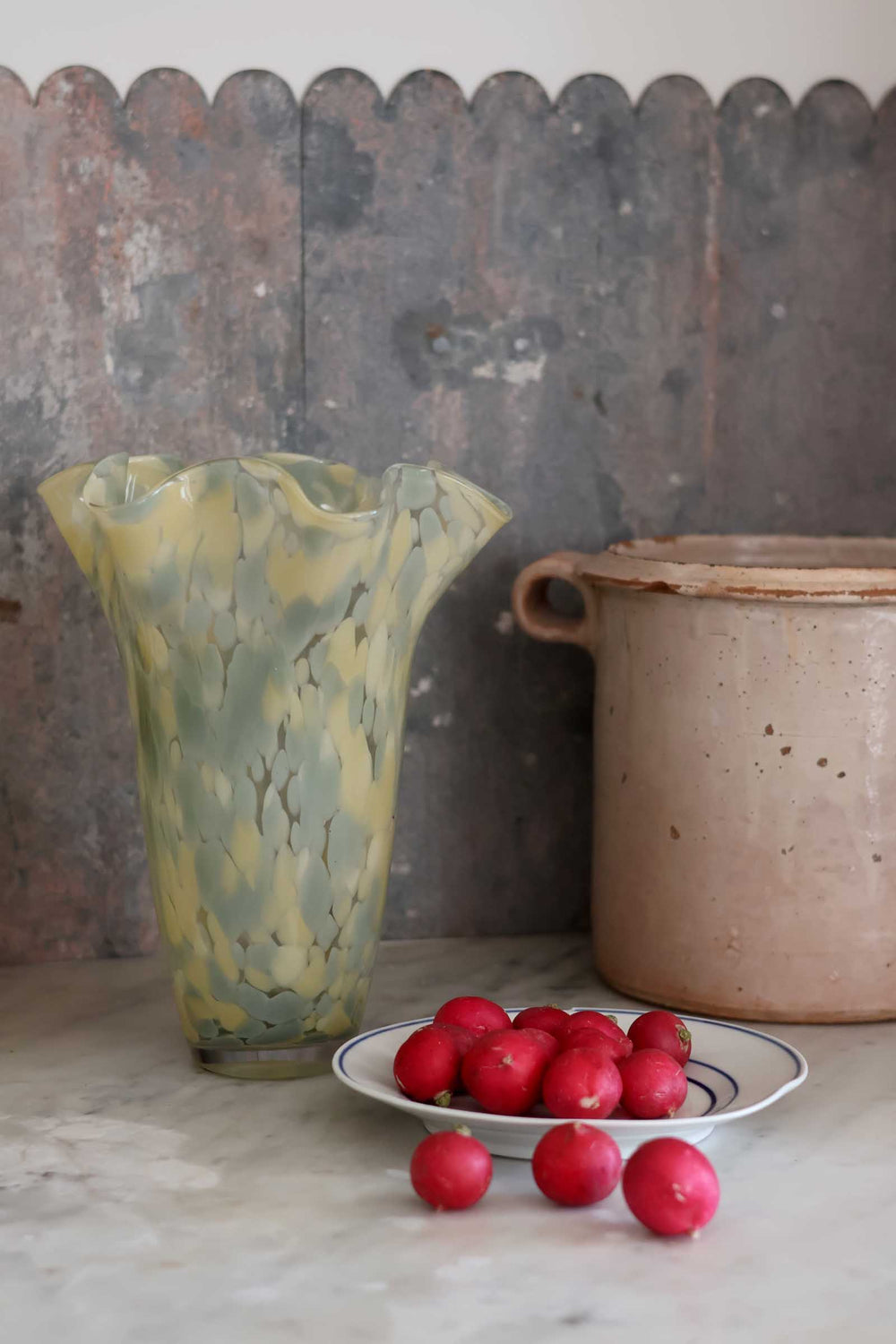 A vintage fluted glass vase with a marbled yellow and green pattern, displayed on a marble surface next to a ceramic pot and a small plate of red berries.