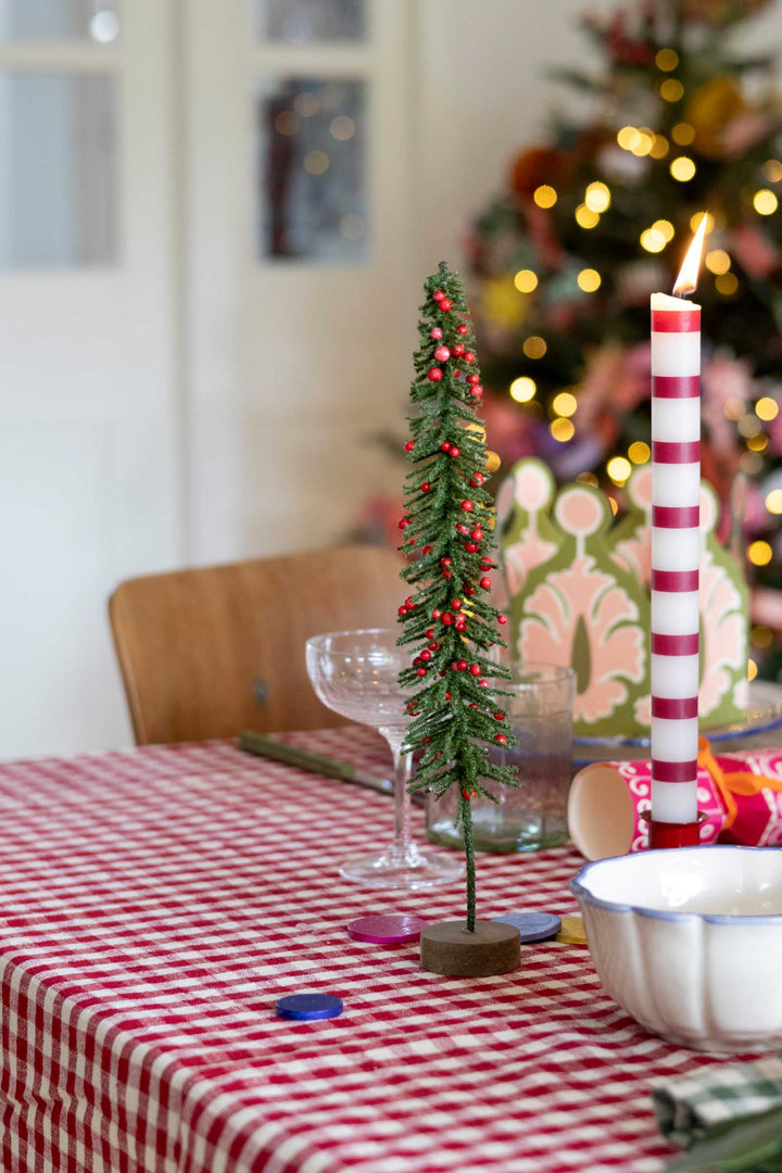 A small decorative Christmas tree with red berries stands on a table covered with a red-and-white checkered tablecloth, alongside a lit red-and-white striped candle, a glass, and a white bowl. A blurred Christmas tree with warm lights appears in the background.