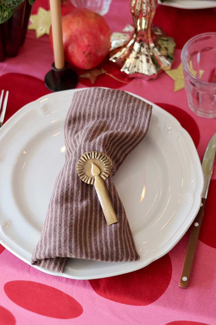 A styled table setting featuring a white ceramic plate with a brown and beige vertically striped linen napkin secured by a gold sunburst ring holder, set on a pink tablecloth with red polka dots.
