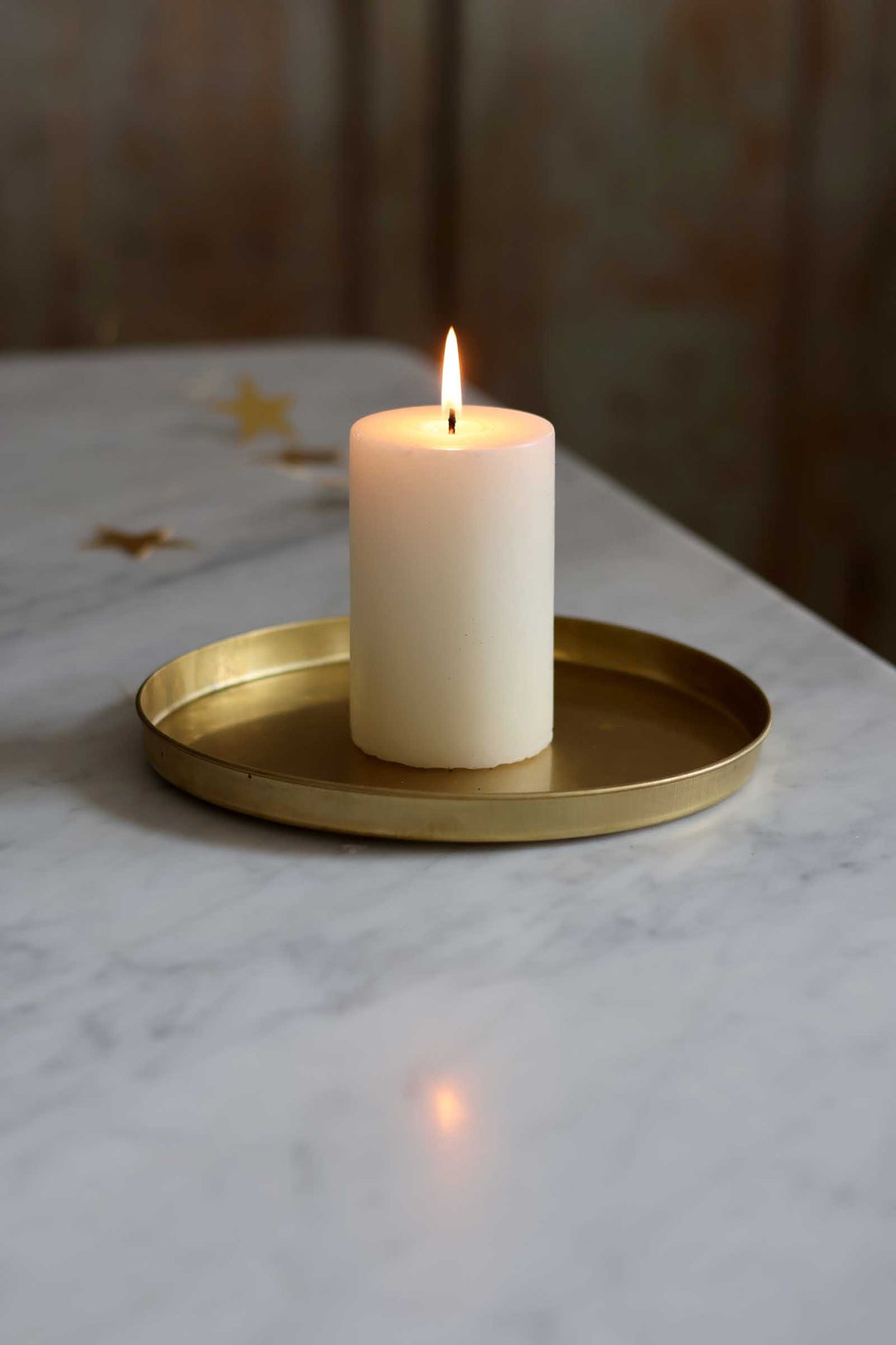 A lit cream-colored pillar candle sits in a round gold metal tray on a white marble surface with a softly blurred brown background.
