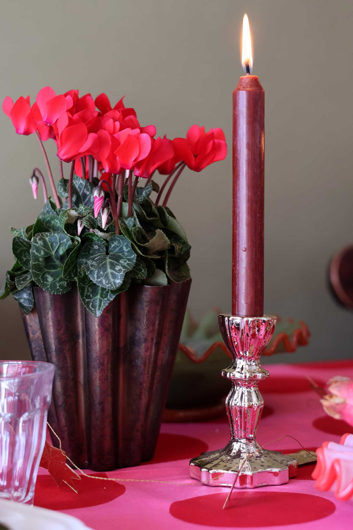 A lit burgundy taper candle in a silver holder stands beside a dark floral arrangement of red cyclamins in a ribbed vase on a pink tablecloth.
