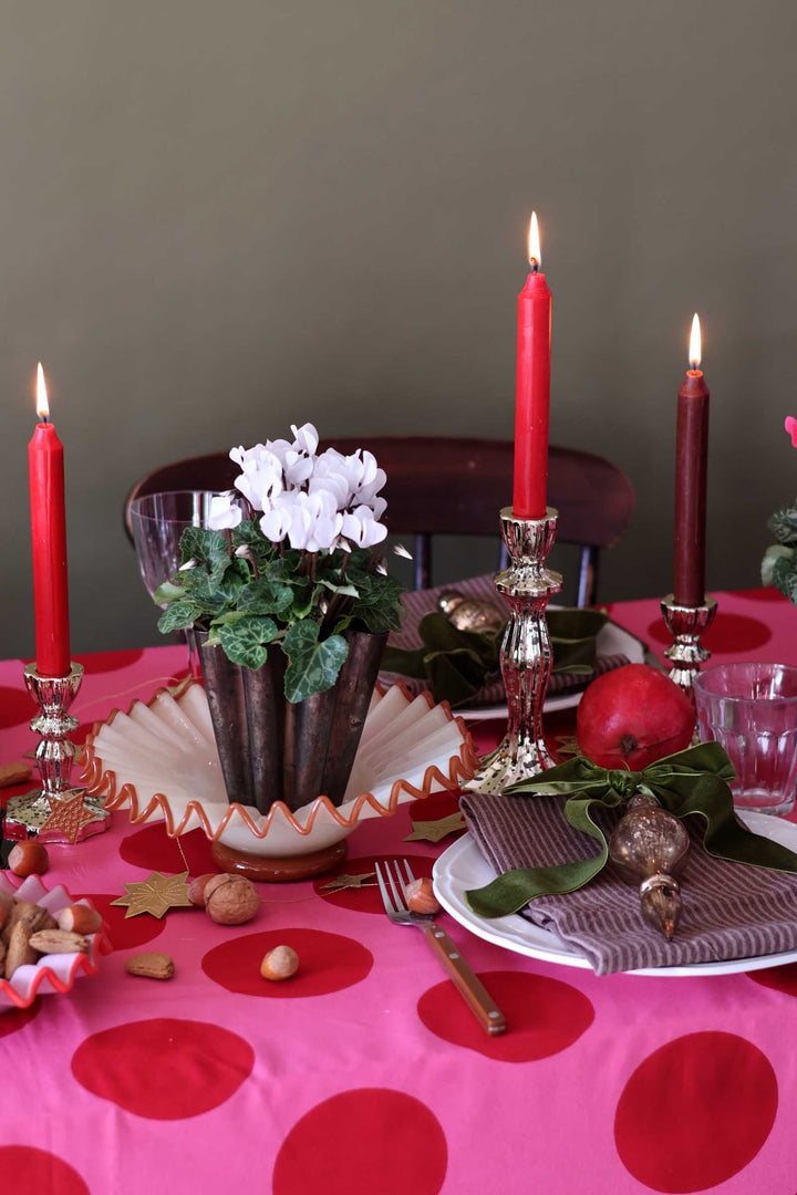 A festive holiday table setting featuring a vibrant pink tablecloth with large red polka dots, decorated with three lit red taper candles in silver candlesticks, a central dark metal vase with white flowers, and a place setting with a striped napkin, a red apple, and green leaves.
