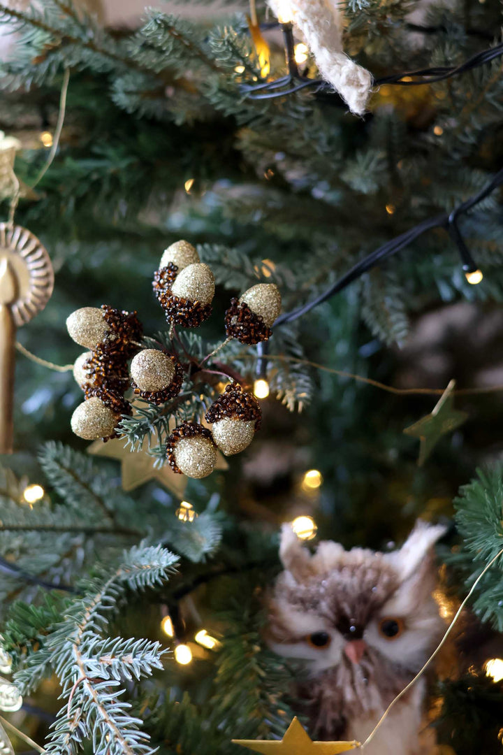 A close-up of a decorated Christmas tree with gold glitter pinecones, warm white string lights, and a fluffy owl ornament peeking out from behind the branches.