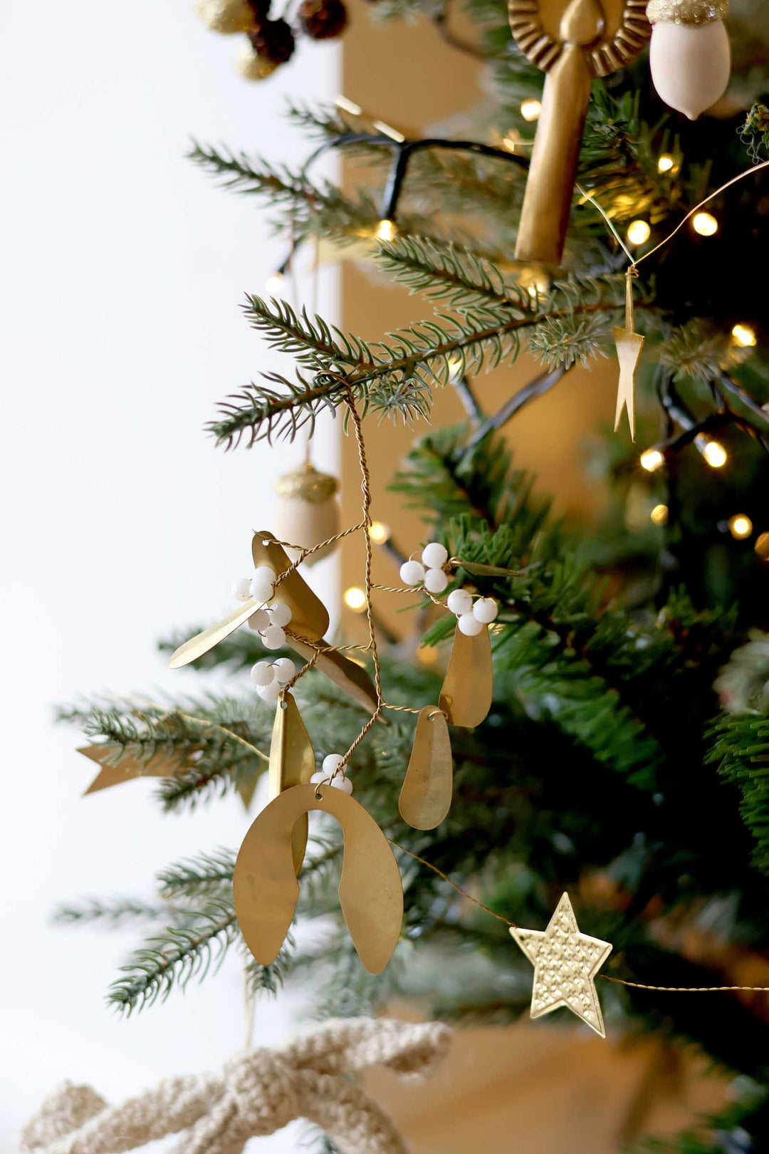Gold mistletoe and star Christmas tree ornaments hanging on a green evergreen tree with warm white string lights.