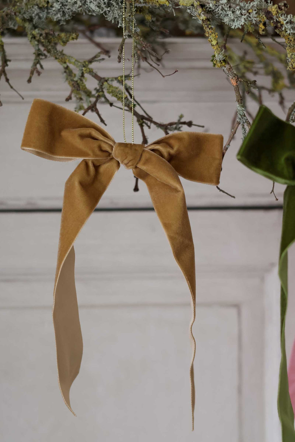 A camel brown velvet bow ornament hanging from a gold chain against a white paneled wall, with dried botanicals and other ornaments partially visible in the background.