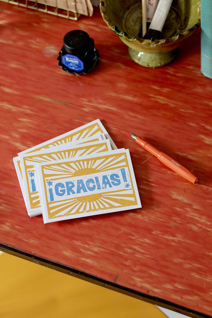 Stack of cards with '¡Gracias!' text on a red surface with a pen and ink bottle.