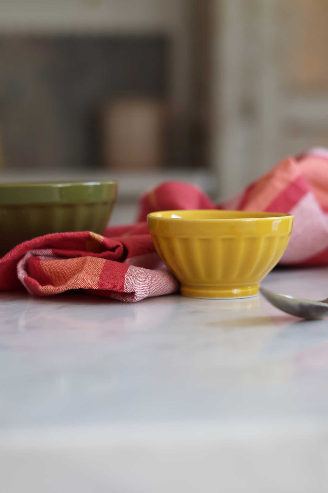 A small yellow ribbed ceramic bowl sits on a light marble countertop next to a red and pink checkered cloth, with another bowl blurred in the background.