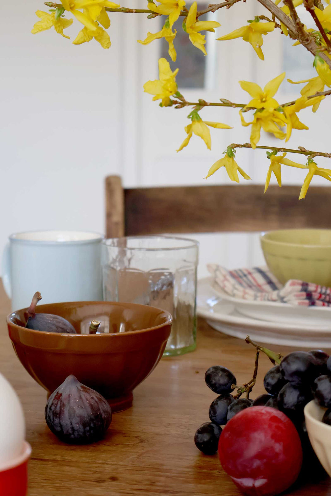 Dining table with fruit, bowls, and a branch of yellow flowers.