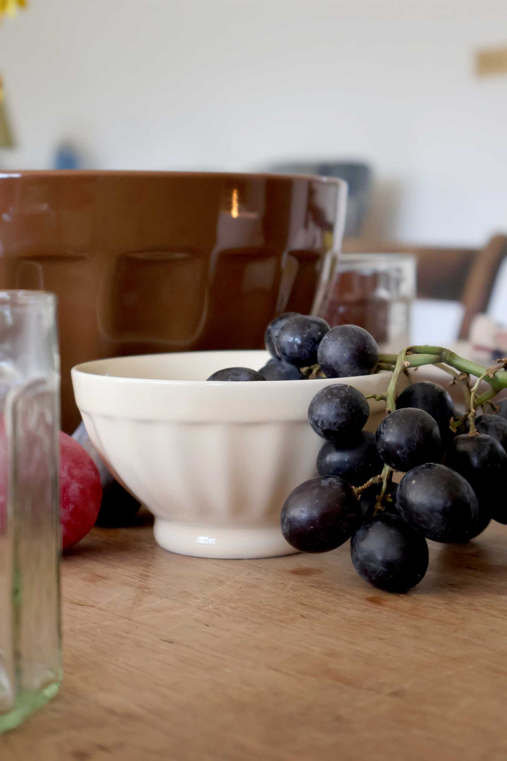 Bunch of grapes on a wooden table with a white bowl and brown pot in the background.
