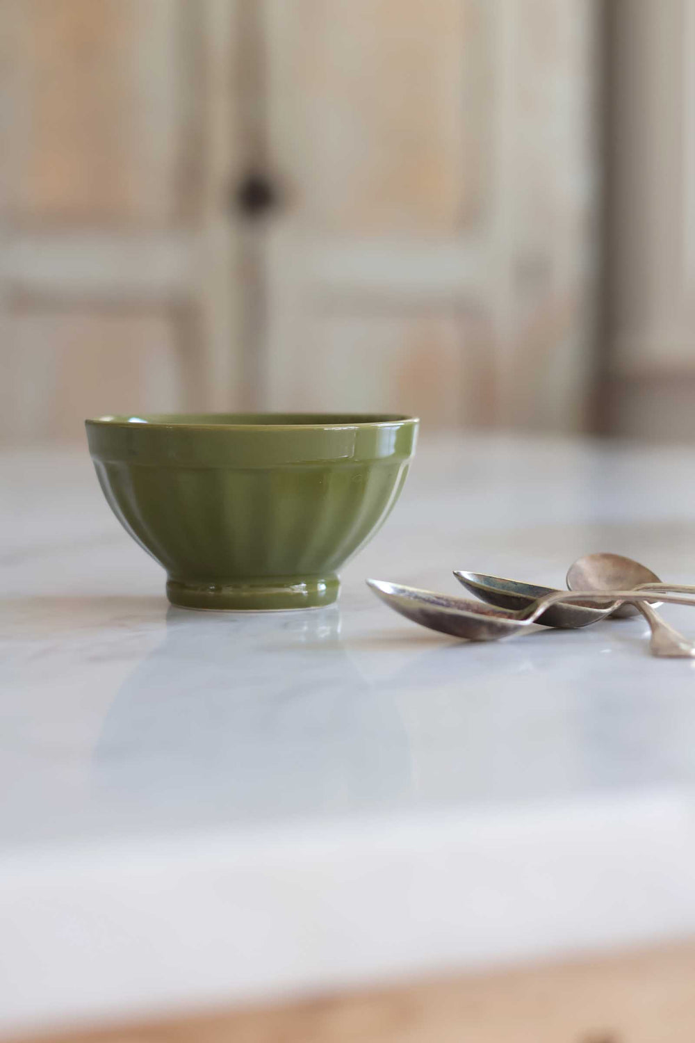 An olive green ceramic bowl with a fluted rim sits on a white marble countertop next to three silver spoons, with a softly blurred kitchen background.