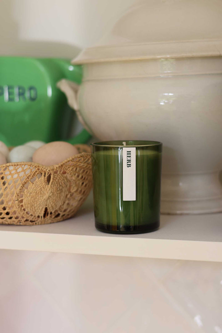 An olive green glass candle jar with a white vertical label reading 'HERB' sits on a white shelf, styled with a woven basket, eggs, and a cream-colored ceramic pot in the background.