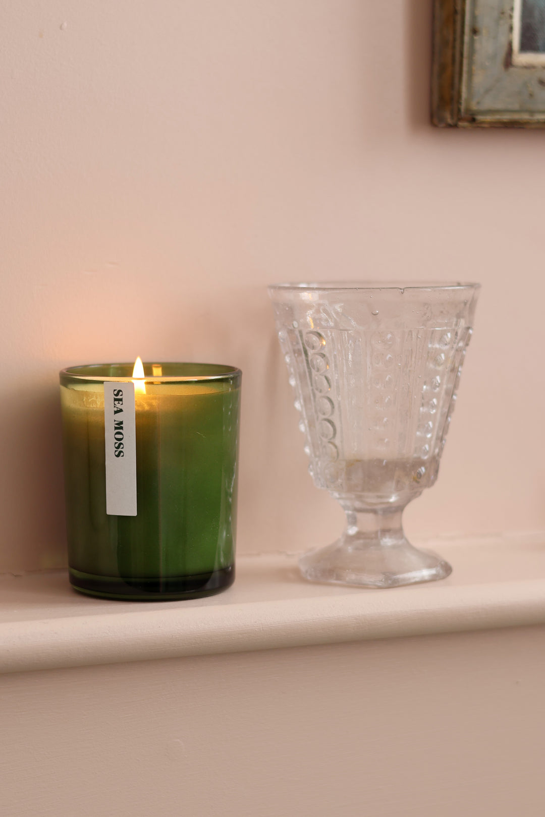 A green glass jar candle with a lit flame and a white vertical label reading 'SEA MOON' sits on a cream-colored windowsill next to a clear textured glass goblet, set against a soft beige wall.