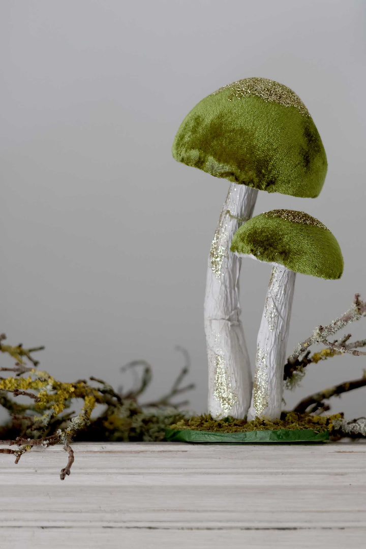 Two decorative velvet mushrooms with olive green caps and white stems, accented with gold glitter, standing on a small green base with faux moss and dried branches.