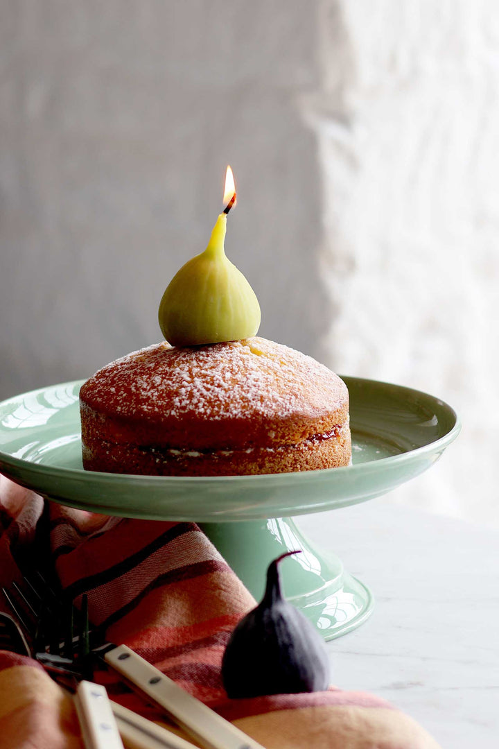 A small layered cake on a pale green pedestal plate, topped with a whole fig and a lit candle. The scene is styled with a fig, wooden utensils, and a patterned cloth against a soft neutral background.