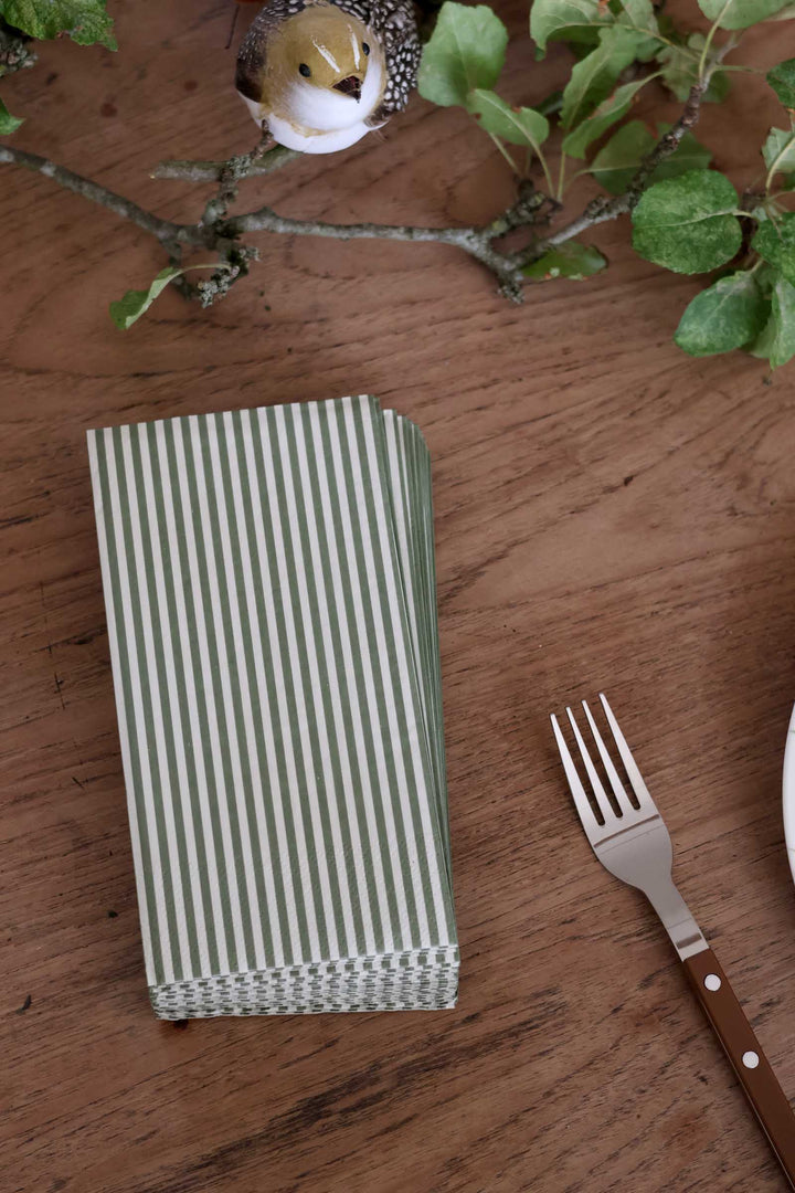 A stack of green and white striped paper napkins on a wooden table, styled with a metal fork, green leaves, and a small bird figurine.
