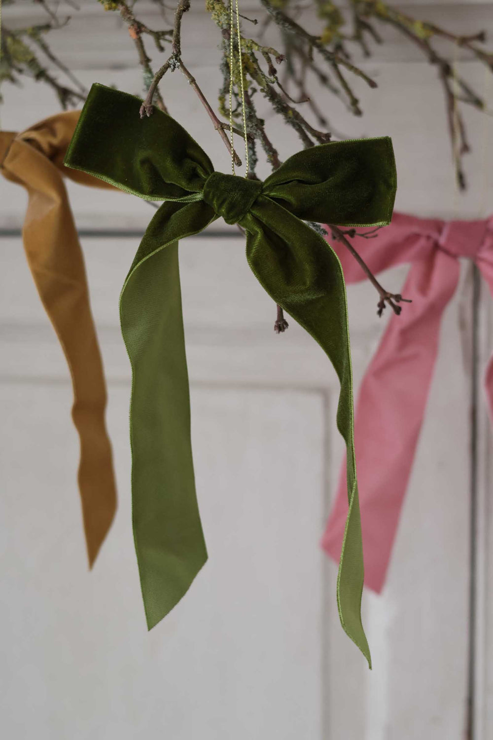 A dark green velvet bow ornament hanging from a branch, with a mustard yellow ribbon on the left and a pink ribbon on the right, set against a soft white paneled background.