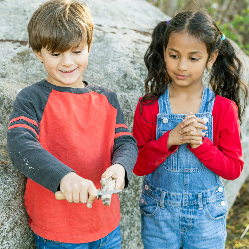 Two children outdoors, one using a carving knife, against a natural rock background.