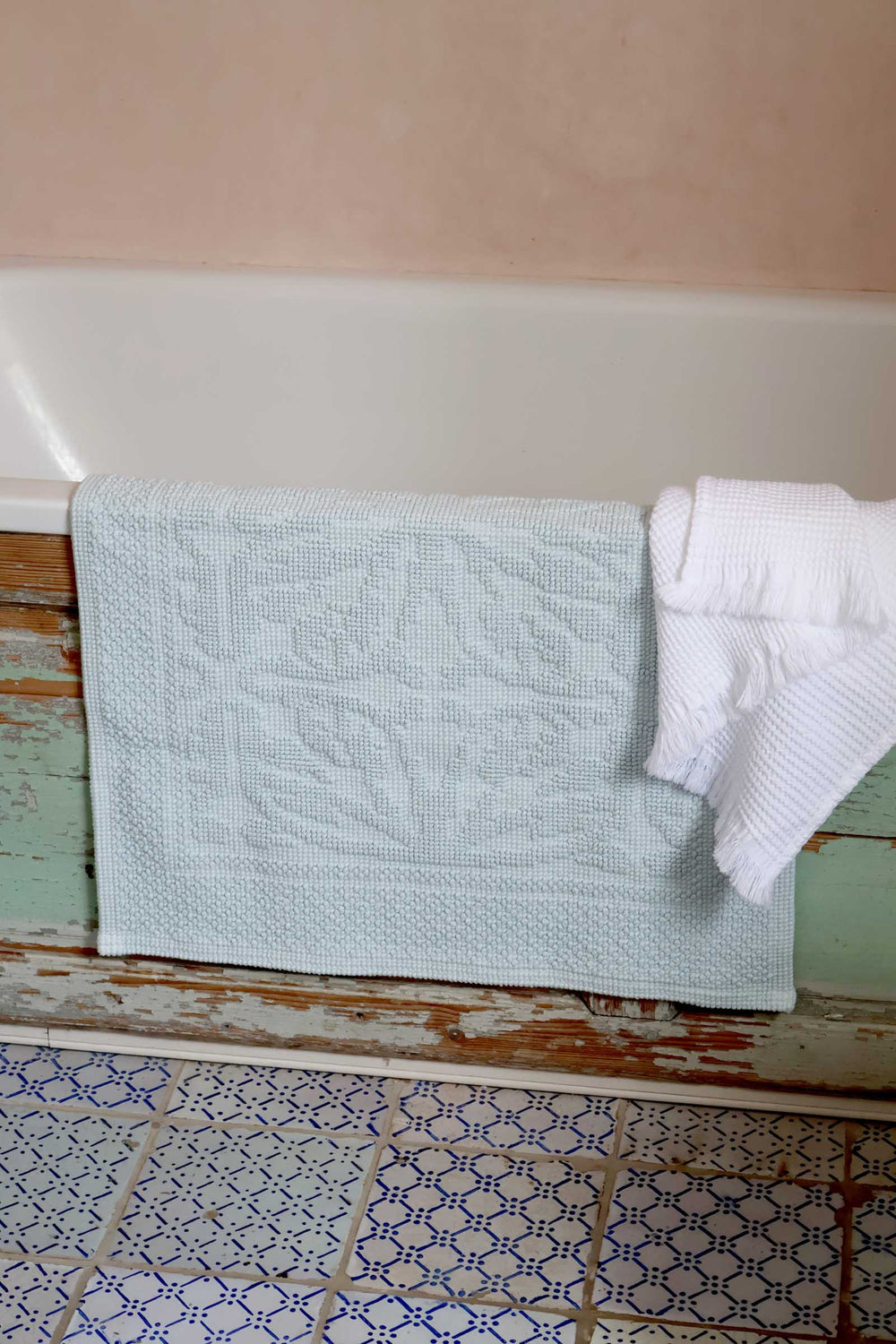 Light blue bathmat draped over a bathtub with a white towel on a wooden ledge.