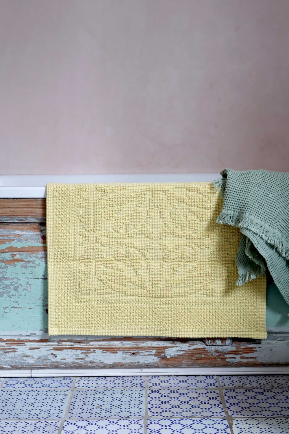Yellow textured bath mat draped over a wooden surface with a pink wall in the background